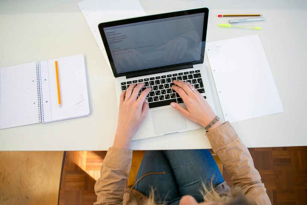 Top view of crop anonymous female student typing on keyboard while preparing for university assignment at table with notebook and papers
